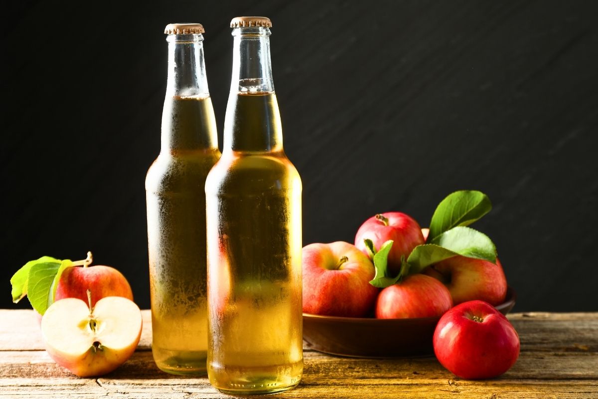 Two glass bottles of vinegar with a bowl of apples on the side.
