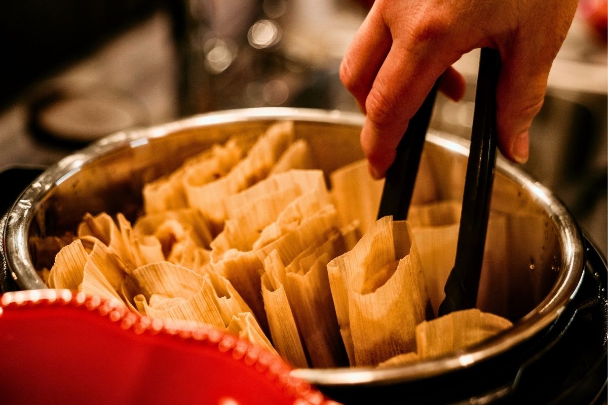 A photo of tamales in a silver bowl with a hand in black tongs picking one.