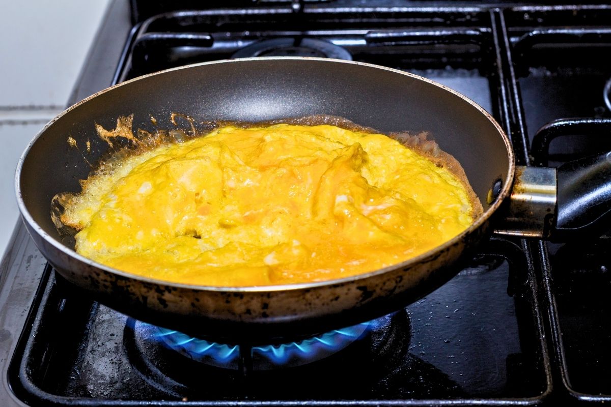 Scrambles eggs coking in a frying pan on a blue gas flame.