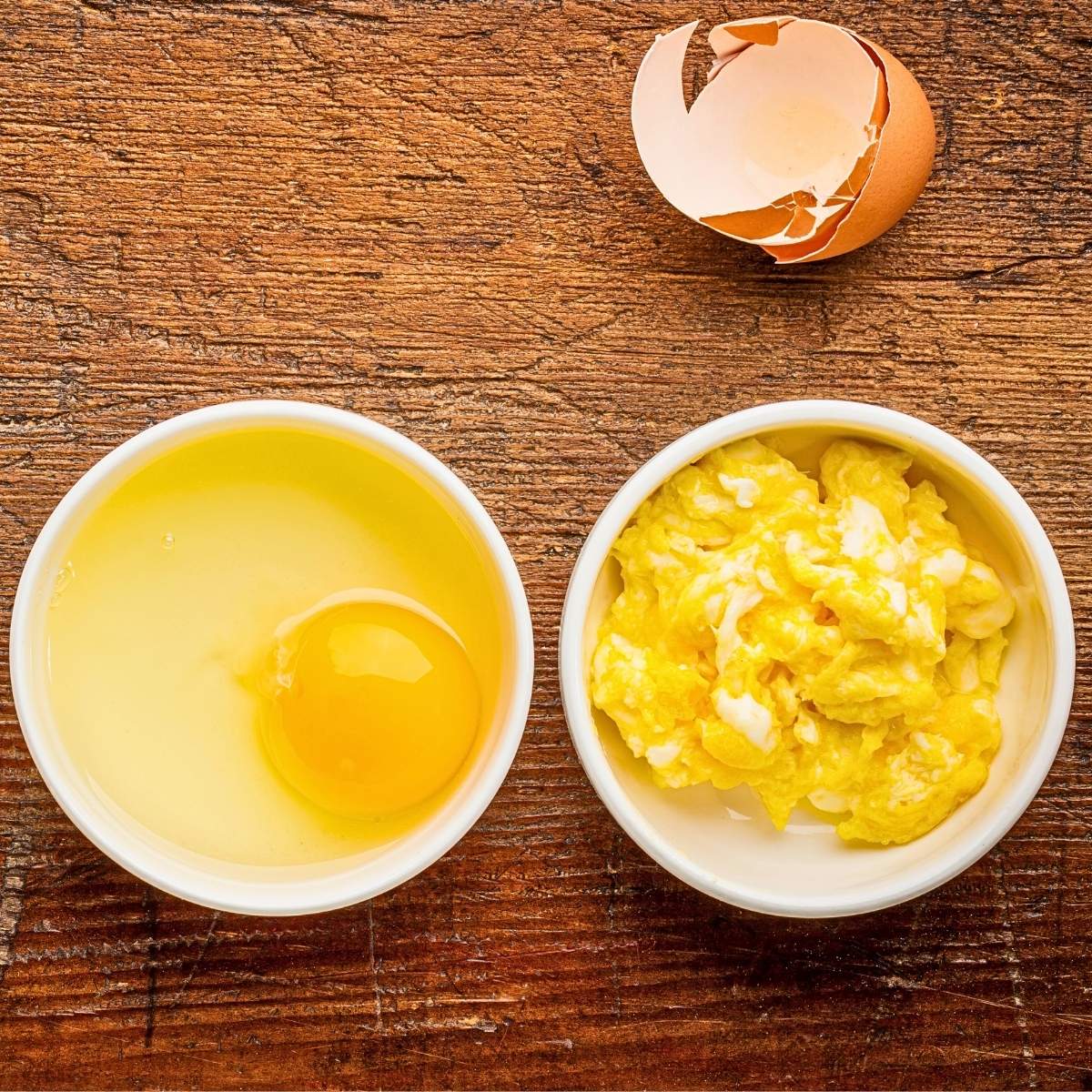 Two white bowls, one with scrambled eggs and another with raw egg. There is an egg shell on the side.