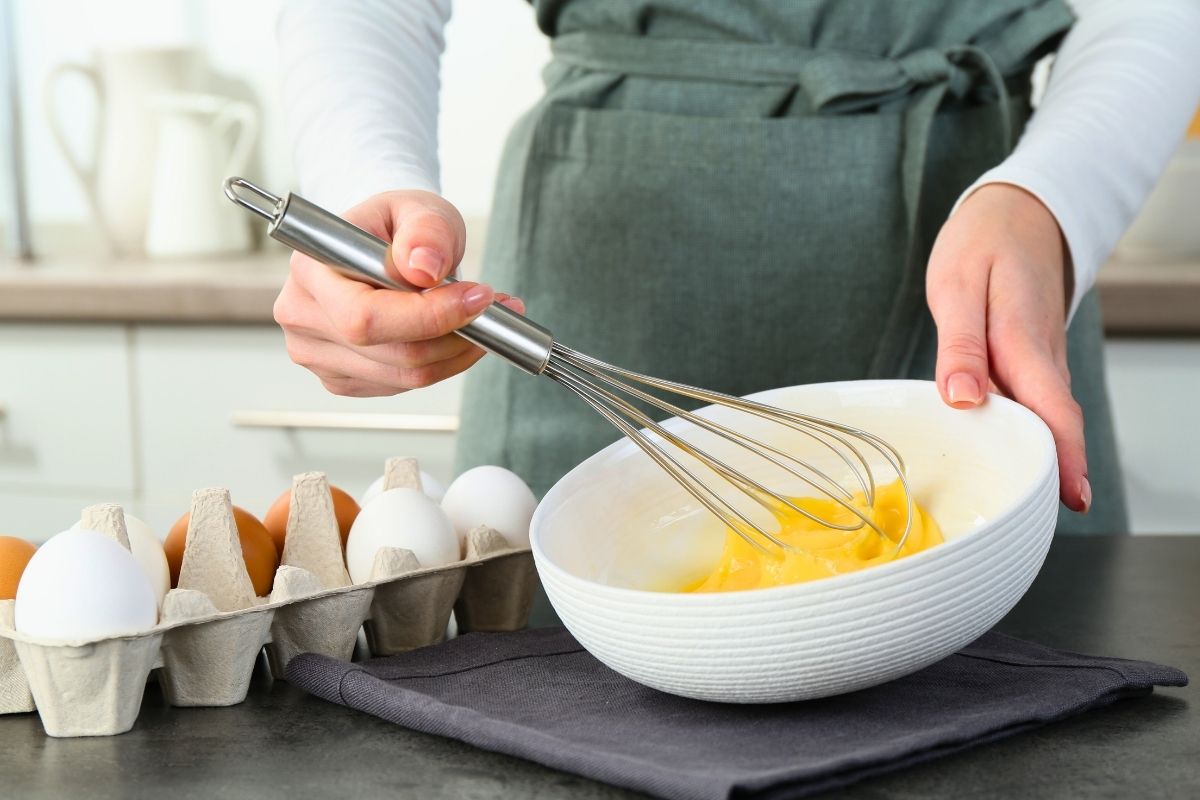 Lady beating eggs with a whisk in a white bowl.